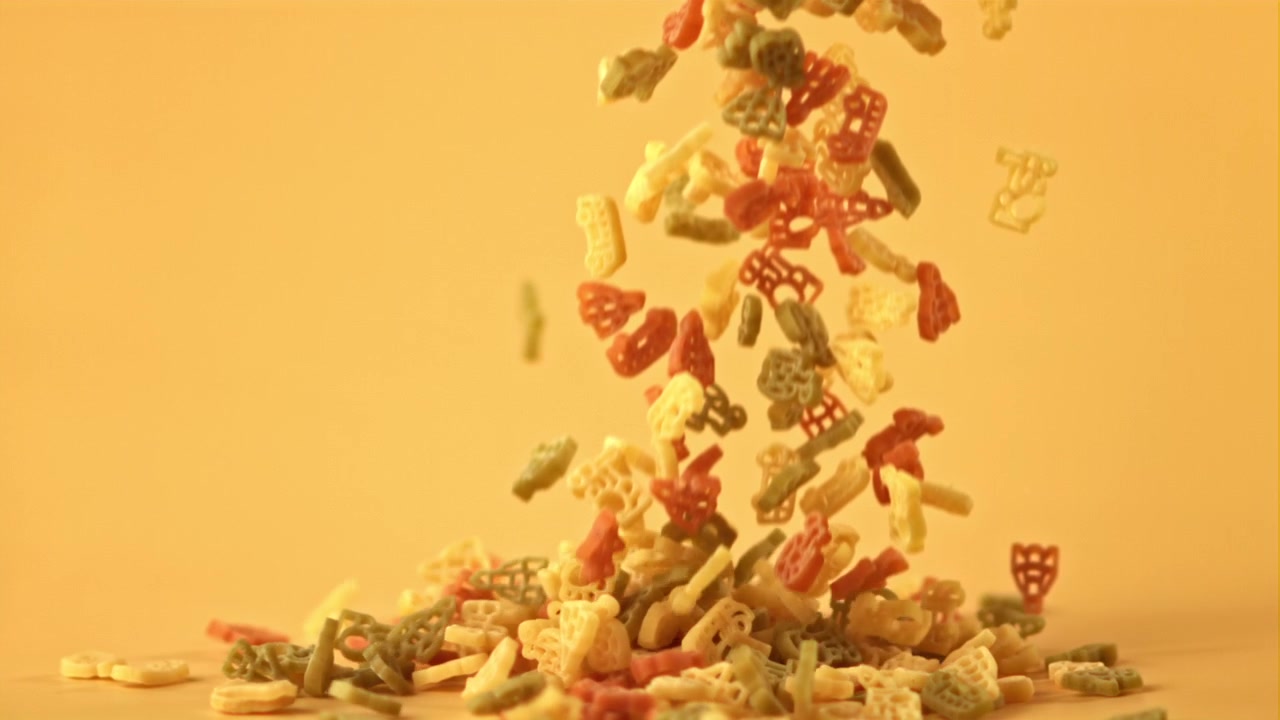 Forme di Pasta Colorate che Cadono al Rallentatore su Sfondo Giallo