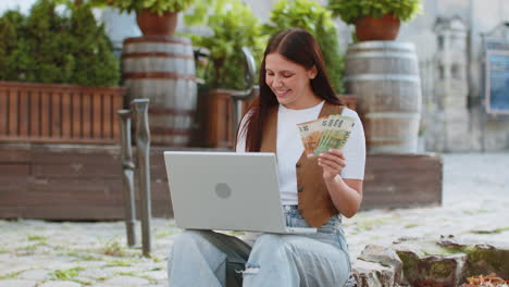 happy rich woman using laptop winner victory holding cash money in euro banknotes on city street