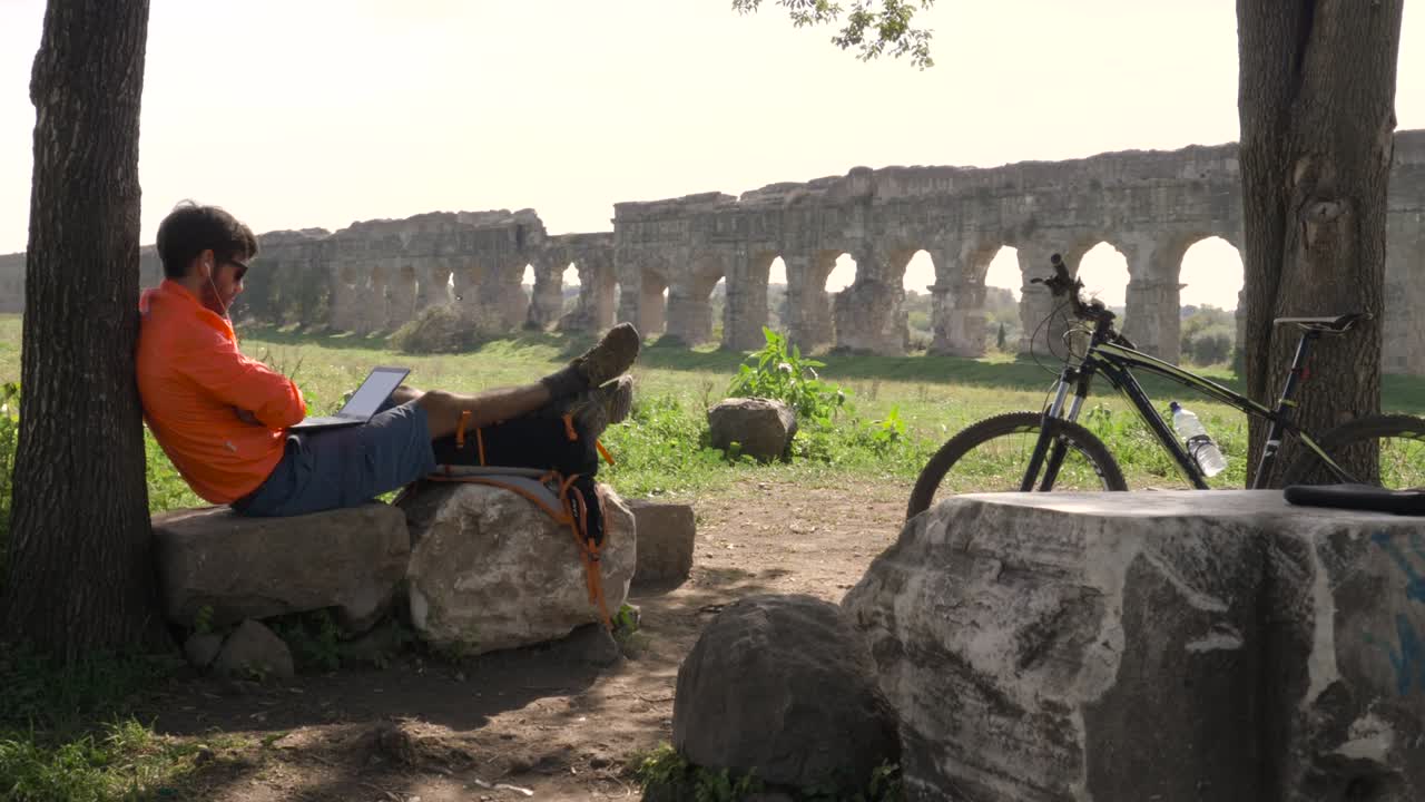 고대 로마의 수로 앞에서 비디오 통화에서 나무 의 바위 위에 앉아있는 자전거 타는 젊은 매력적인 운동 선수, 오렌지색 스포츠 의류와 배, 로마의 parco degli acquedotti