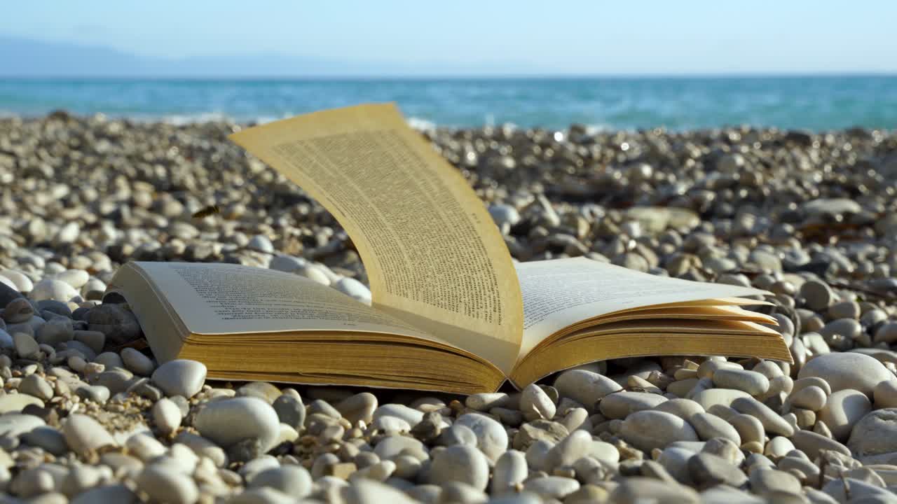 Pagine di libro aperte dal vento su una spiaggia con ciottoli e onde del mare sullo sfondo, svago in vacanza