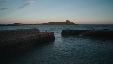 a man walks his dog to the end of a stone pier at colliemore harbour, dublin, with dalkey island in the background. the sea is moderate with gentle waves under a soft evening sky.