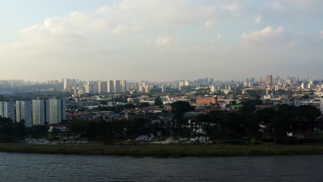 Aerial-drone-left-trucking-and-panning-shot-of-the-south-of-São-Paulo,-Brazil-from-the-man-made-Guarapiranga-Reservoir-towards-the-Socorro-neighborhood-with-skyscrapers-and-houses-during-sunset