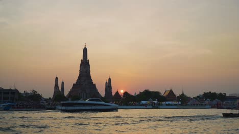 Fascinante-Plano-General-Del-Templo-Wat-Arun-En-Bangkok-Durante-Una-Impresionante-Puesta-De-Sol,-Con-Barcos-Cruzando-Frente-Al-Majestuoso-Río-Chao-Phraya