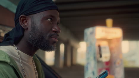 african american man scrolling on phone with smile by car under bridge