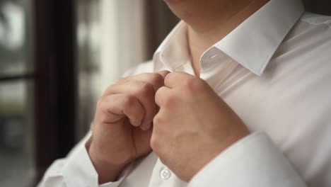 man buttons up his white shirt standing in the front of a bright window 02