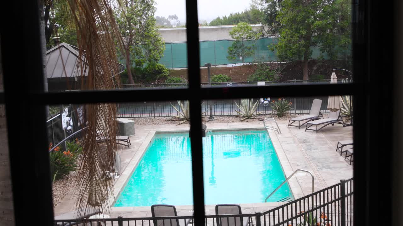 Hand moving into frame to pull back a curtain in a hotel room to reveal a bright blue pool and palm trees