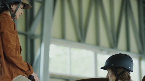 two women wearing riding helmets interacting with a horse in an indoor arena