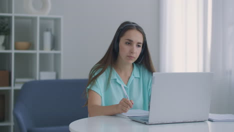 Studentin-Sitzt-Zu-Hause-Am-Schreibtisch-Und-Trägt-Ein-Headset,-Spricht-Aus-Der-Ferne-Per-Videoanruf-App-Und-Laptop-Gerät.-E-Learning-Sprache,-Lernen-Mit-Einem-Tutor-Oder-Klassenkameraden,-Erledigt-Gemeinsame-Aufgaben-Aus-Der-Ferne.-Modernes-Technologiekonzept