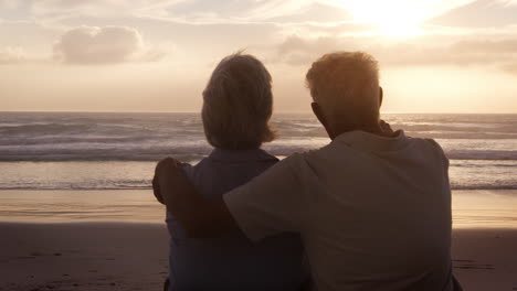 vista trasera de una pareja de ancianos en la playa viendo la puesta de sol sobre el océano