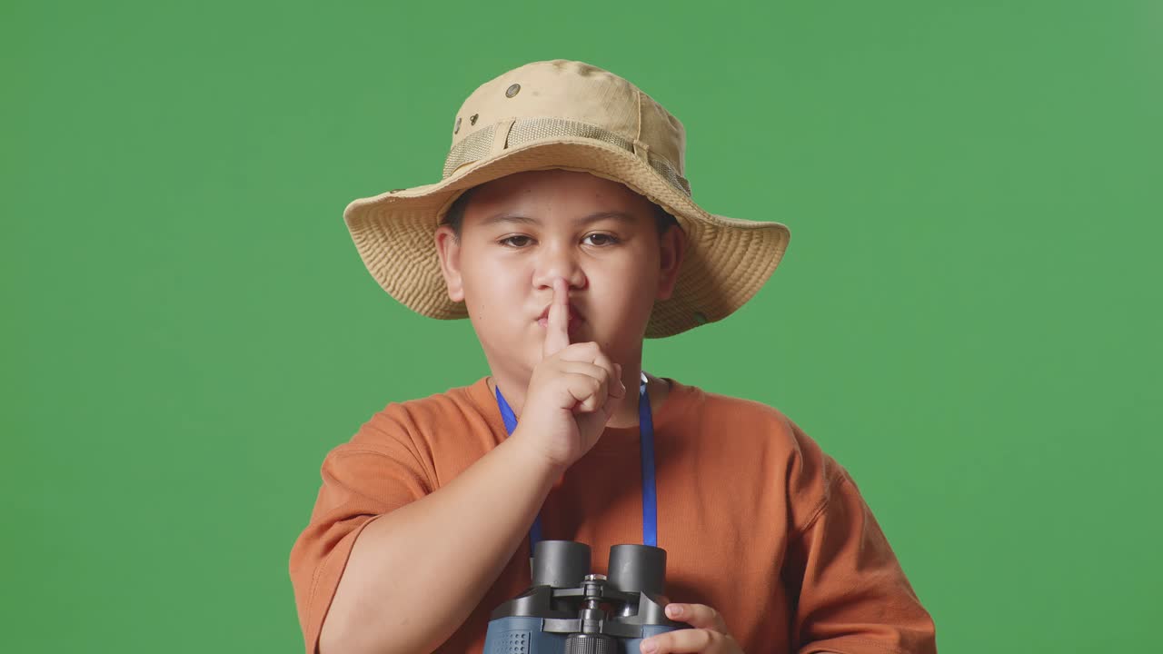 모자를 입은 아시아 관광 소년은 안경을 통해보고 녹색 스크린 배경에 shh 제스처를합니다. 소년 연구자는 무언가를 조사합니다. 여행 관광 모험 개념, 근접