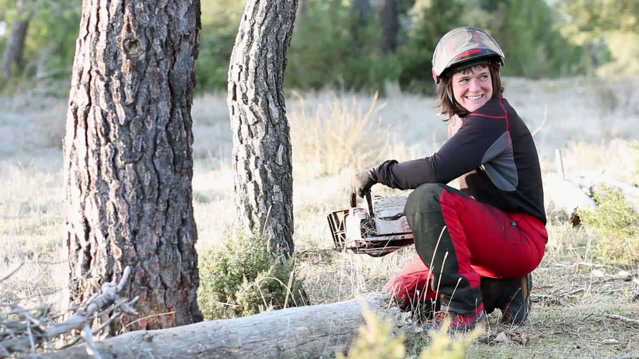 mujer joven sonriendo a la cámara equipada con una motosierra, guantes y casco rojo