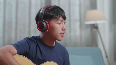close up of asian boy composer with notebook on table wearing headphones and smiling to camera while playing guitar at home