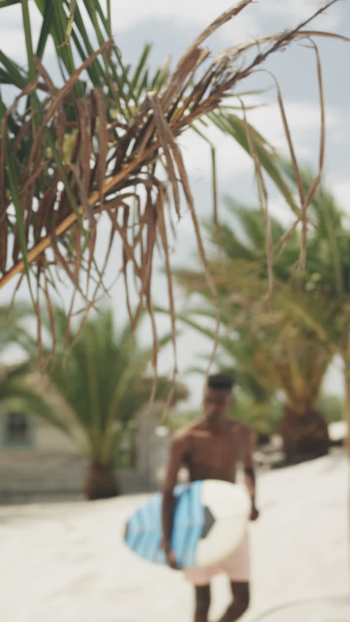 Vertical video of african american man carrying surfboard walking on sunny beach, slow motion