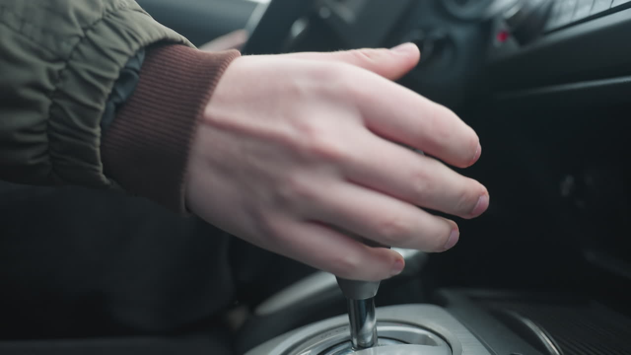 Close up of person pulling gear shift lever back while hand rests on wheel showing smooth manual transition between drive and park modes in car center console control interior under ambient light