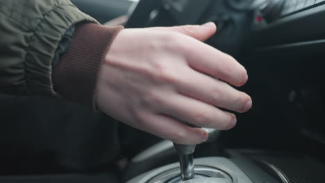 close up of person pulling gear shift lever back while hand rests on wheel showing smooth manual transition between drive and park modes in car center console control interior under ambient light