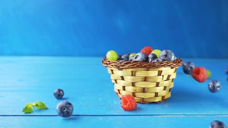 wild berries are pouring out of the falling basket. a basket with different berries falling on a wooden blue table. 4k slow motion video with speed ramp effect.