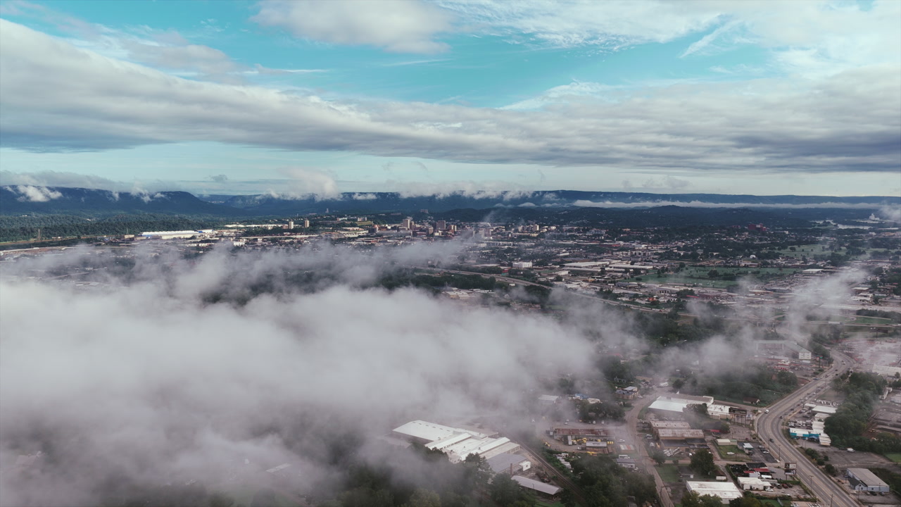 Aerial drone view of Chattanooga shows the city wrapped in drifting low clouds, with mountain ridges framing the skyline under a bright blue sky.