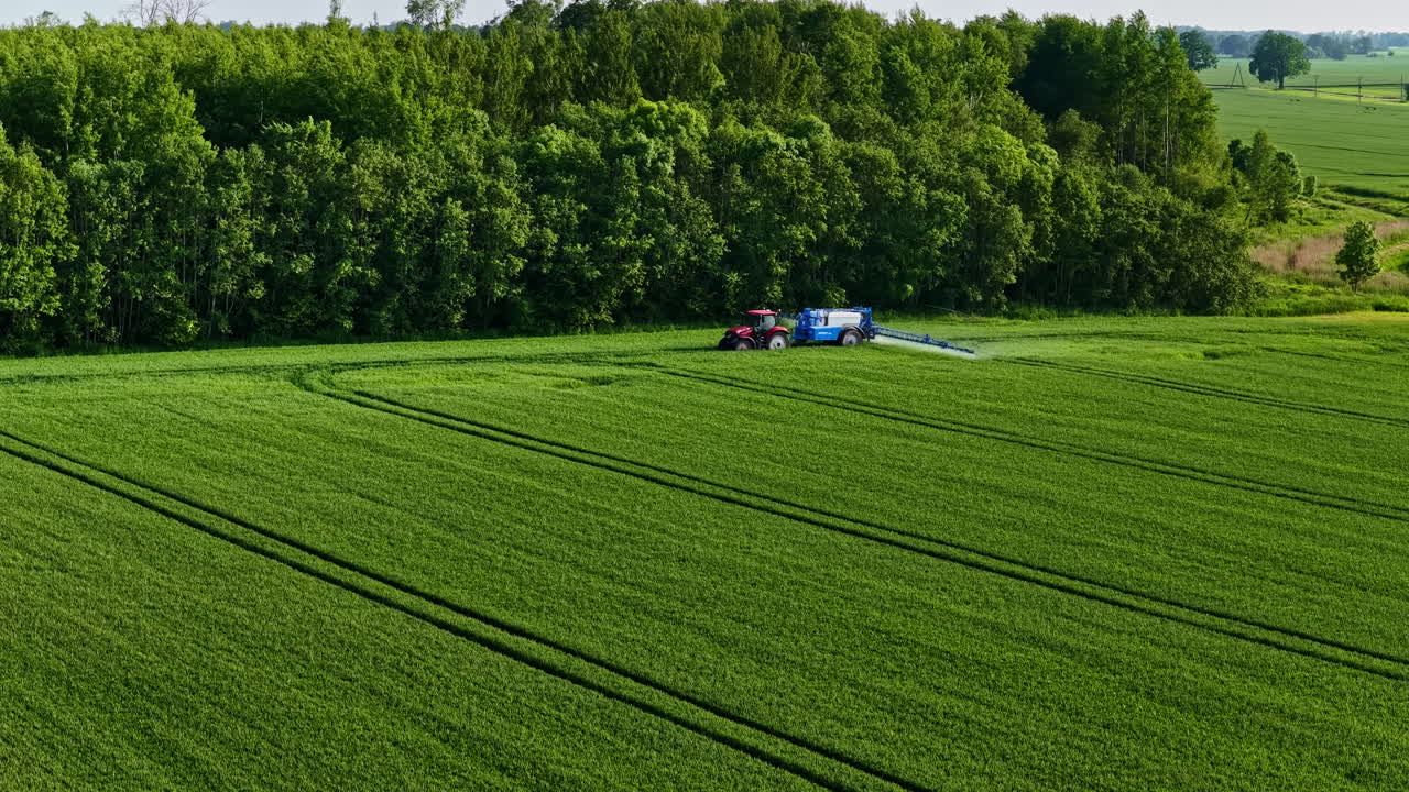 A red tractor sprays crops in a lush green field bordered by dense forest in Latvia, illustrating modern summer agriculture from above