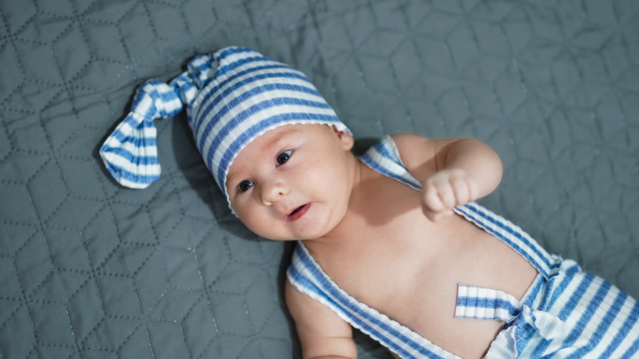 Small kid in gnome costume left on the bed. Babe in striped funny hat turning his head. Close up. Grey background.