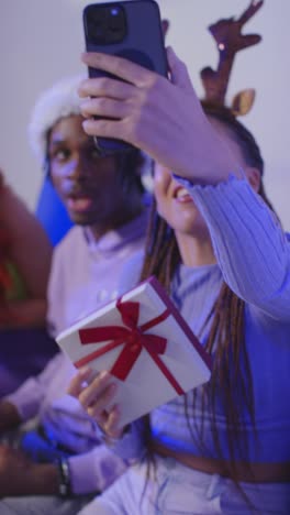 Foto-De-Estudio-De-Vídeo-Vertical-De-Amigos-De-La-Generación-Z-En-Navidad-Sentados-En-Un-Sofá-Con-Gorro-De-Papá-Noel-Y-Astas-De-Reno-Tomándose-Un-Selfie-En-Un-Teléfono-Móvil