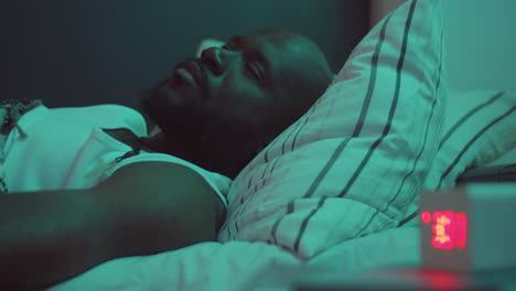 sleepless african american man lying on bed and looking at clock in night