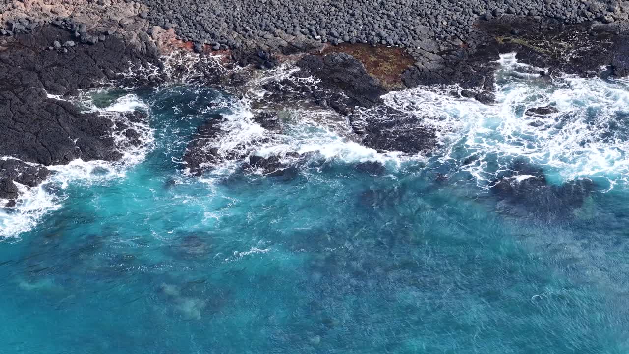 Drone riprende onde turchesi che si infrangono su rocce vulcaniche scure, luce diurna intensa, prospettiva aerea stabile