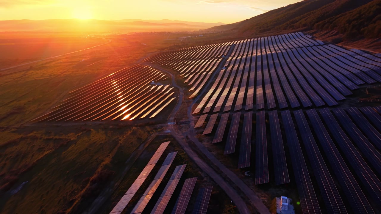 Solnedgangsglød reflekteres på solcelleparkens bjergside, Bulgarien. Droneudsigt over omfattende panelarray med sol, der reflekteres fra modulerne, orange horisont, bjergsilhuet og landlig dal nedenfor.
