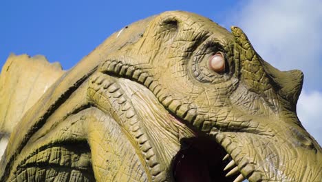 primer plano de ojos aterrador modelo realista gigante de dinosaurio alejándose revelando su cuerpo boca grande dientes aterradores escena de película de suspenso cielo azul nubes cámara lenta