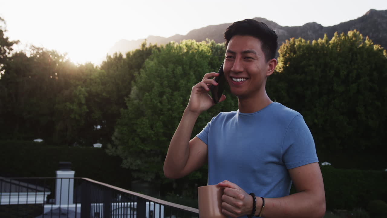 Asian man enjoying phone call on balcony, holding coffee mug, smiling warmly, copy space