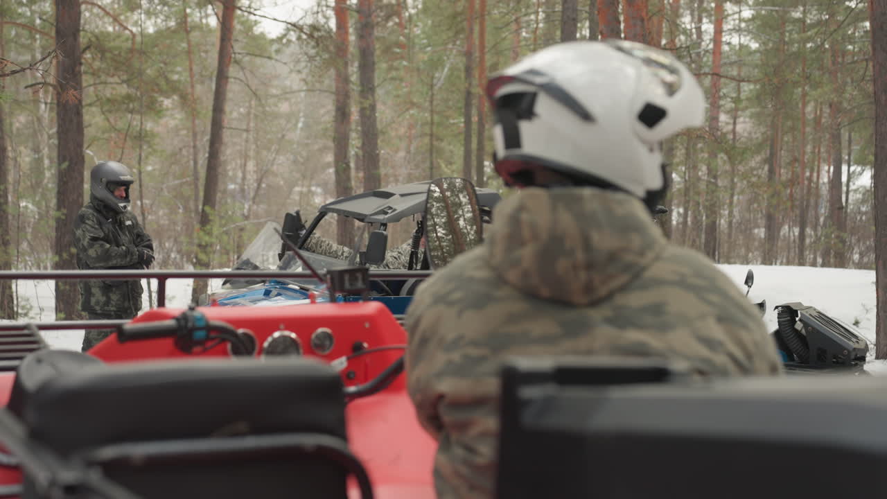Pilota di ATV rosso segue il convoglio attraverso un sentiero innevato di pini, il pilota con casco afferra il manubrio mentre osserva un UTV blu e l'equipaggio in mimetica, i motori rombano e il sibilo dell'acceleratore, il vento freddo e il cielo coperto accentuano.