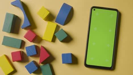 Overhead-Shot-Of-Colourful-Wooden-Building-Blocks-On-Yellow-Studio-Background-With-Green-Screen-Mobile-Phone-1