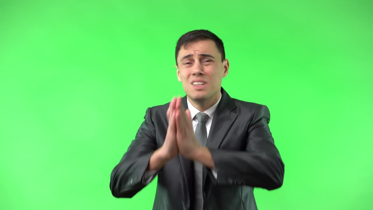 A man in a suit pleads desperately with clasped hands against a green screen