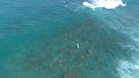Aerial-view-of-surfers-swimming-out-to-sea-over-the-shallow-waters-of-the-reef-in-Cabo,-Mexico