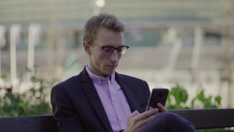 Thoughtful-young-man-sitting-on-bench-and-using-smartphone