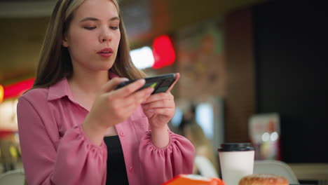 woman in pink dress focusing intently on her phone, adjusting to take a picture of something in front of her, coffee cup and burger in the foreground on the table, with a softly blurred background