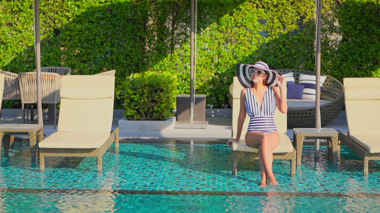 Regal Asian woman suntanning next to the pool sitting on a deckchair at royal tropical resort wearing stripped monokini and sunhat daytime
