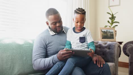 Happy-african-american-father-sitting-on-sofa-with-son-on-lap-using-tablet,-in-slow-motion