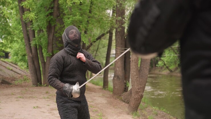 Close up of man practicing sword fighting in medieval armor with another man demonstrating various stances and movements. scene features detailed black padded clothing and metal fencing