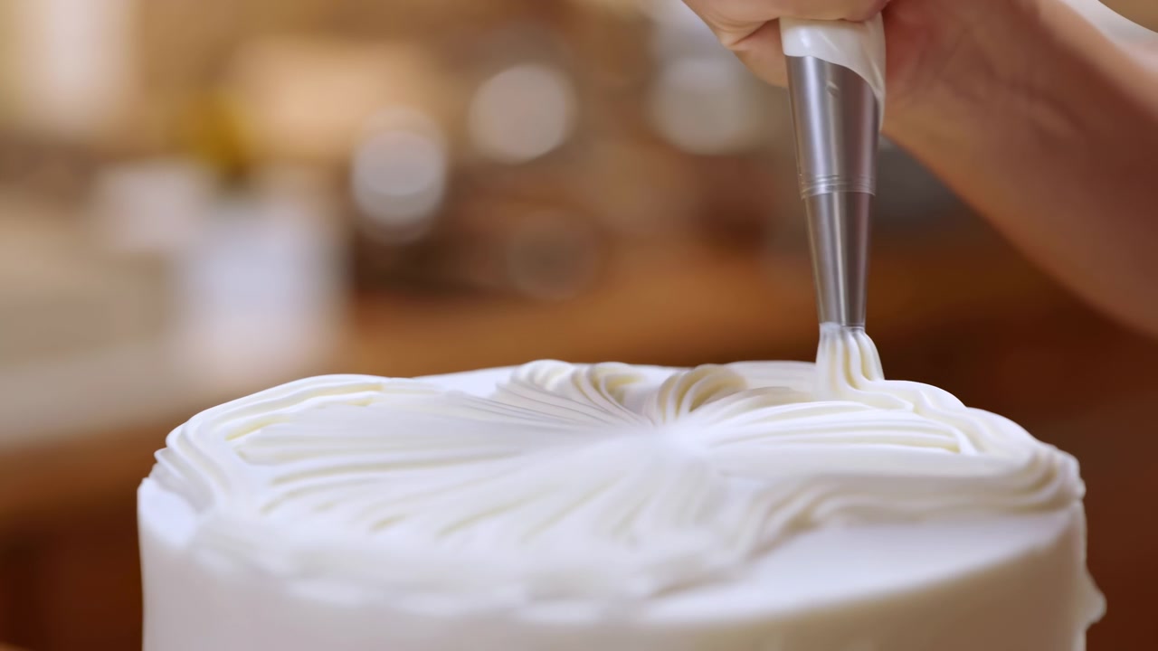 Hand steadies piping bag with metal tip while pressing white buttercream onto round cake, forming defined ridged swirl in warm kitchen light with soft blur behind, showing careful dessert decoration
