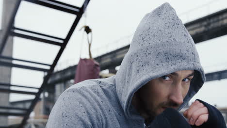 close-up view of handsome bearded caucasian man in grey hoodie and boxing to the camera outdoors an abandoned factory on a cloudy morning