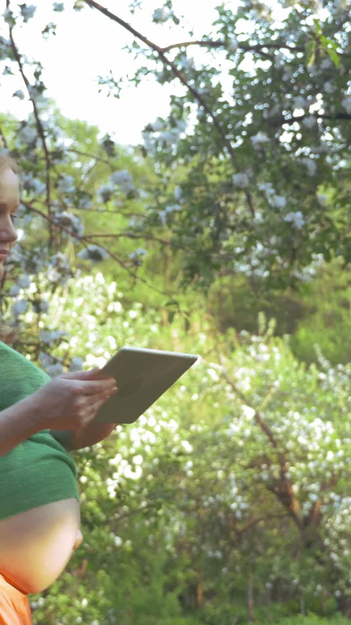 Pregnant woman using a tablet in a blossoming garden