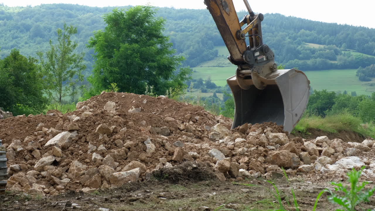 Excavator bucket digging into a pile of dirt and rocks Trees and green hills are in the background