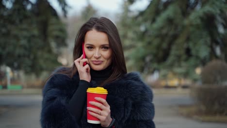 pretty woman with a phone and a cup of hot coffee or tea. portrait of a beautiful girl in luxury fur coat talking the mobile phone and drinking coffee in the park.