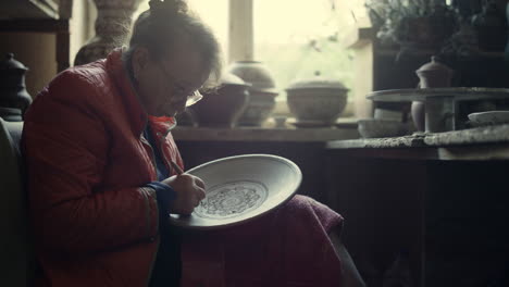 elderly woman making ornament on wet product in studio. ceramist scraping plate