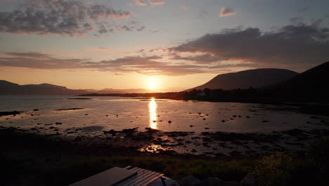 aerial low flyover shot of a camper van on the shore of a calm peaceful sea with the sun setting among hills and clouds