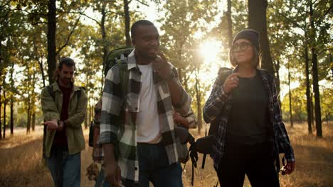 Happy-international-group-on-a-hike-in-the-summer-forest.-A-man-with-Black-skin-in-a-checkered-shirt-and-a-girl-with-a-bob-with-blond-hair-walk-through-the-forest-with-their-friends-in-special-hiking-clothes-in-the-summer