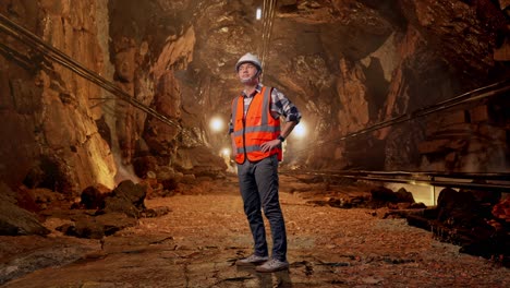 full body side view of asian male engineer wearing safety helmet looking around while standing with arms akimbo in underground mine tunnel