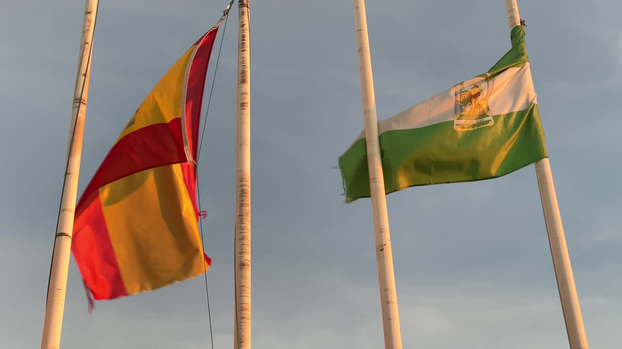 a Spanish and Andalusian flags flap in the warm evening breeze above the beach of Sanlucar de Barrameda under golden sky light
