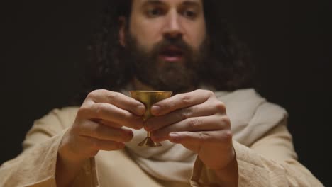 Foto-De-Estudio-De-Un-Hombre-Vestido-Con-Túnicas-Con-Cabello-Largo-Y-Barba-Que-Representa-La-Figura-De-Jesucristo-Bendiciendo-El-Cáliz-Del-Vino