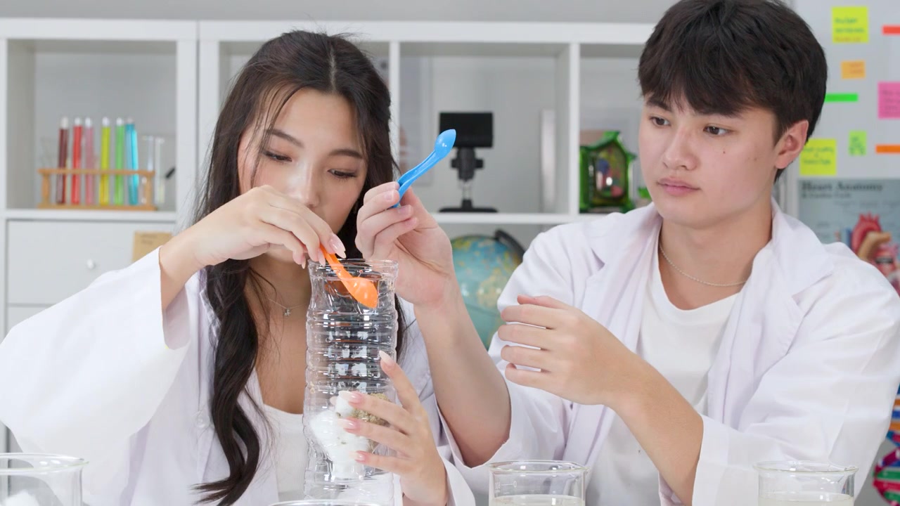 นักเรียนสองคนในชุดเสื้อกาวน์กำลังสร้างเครื่องกรองน้ำ DIY โดยใช้ขวดพลาสติก